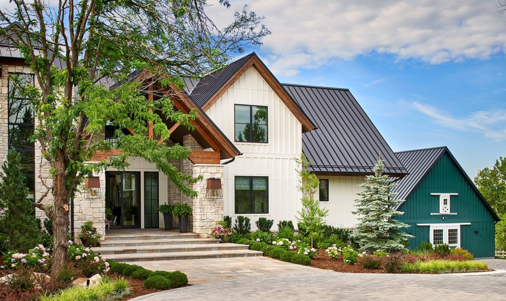 Modern house with a pitched roof, stone and wood facade, surrounded by landscaped greenery, under a partly cloudy sky.