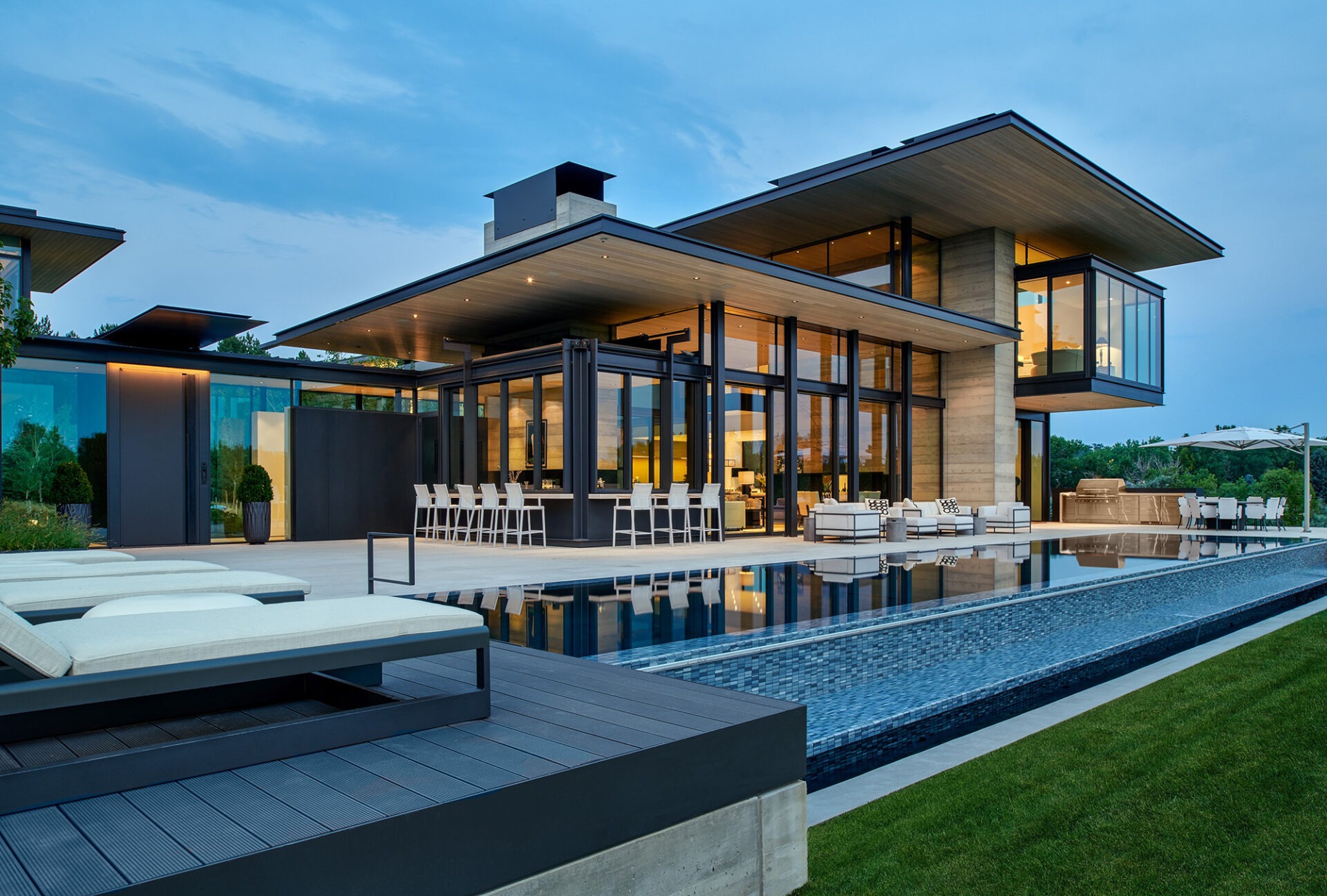 Modern house with large glass windows, expansive terrace, sleek pool, and outdoor seating area. Evening setting with lush greenery and serene sky backdrop.