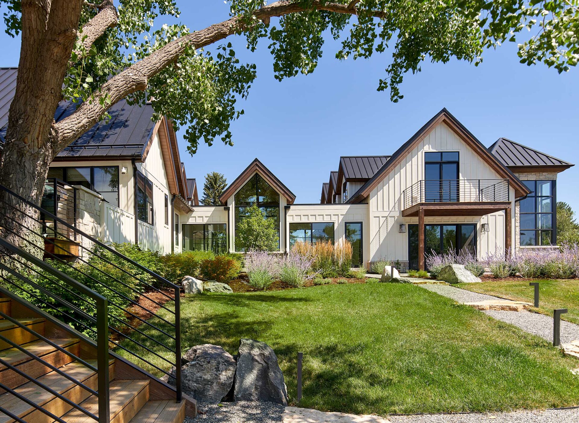 Modern house with a gabled roof amidst greenery, pathways, and trees. Large windows and sleek design create an inviting, spacious appearance under a clear sky.