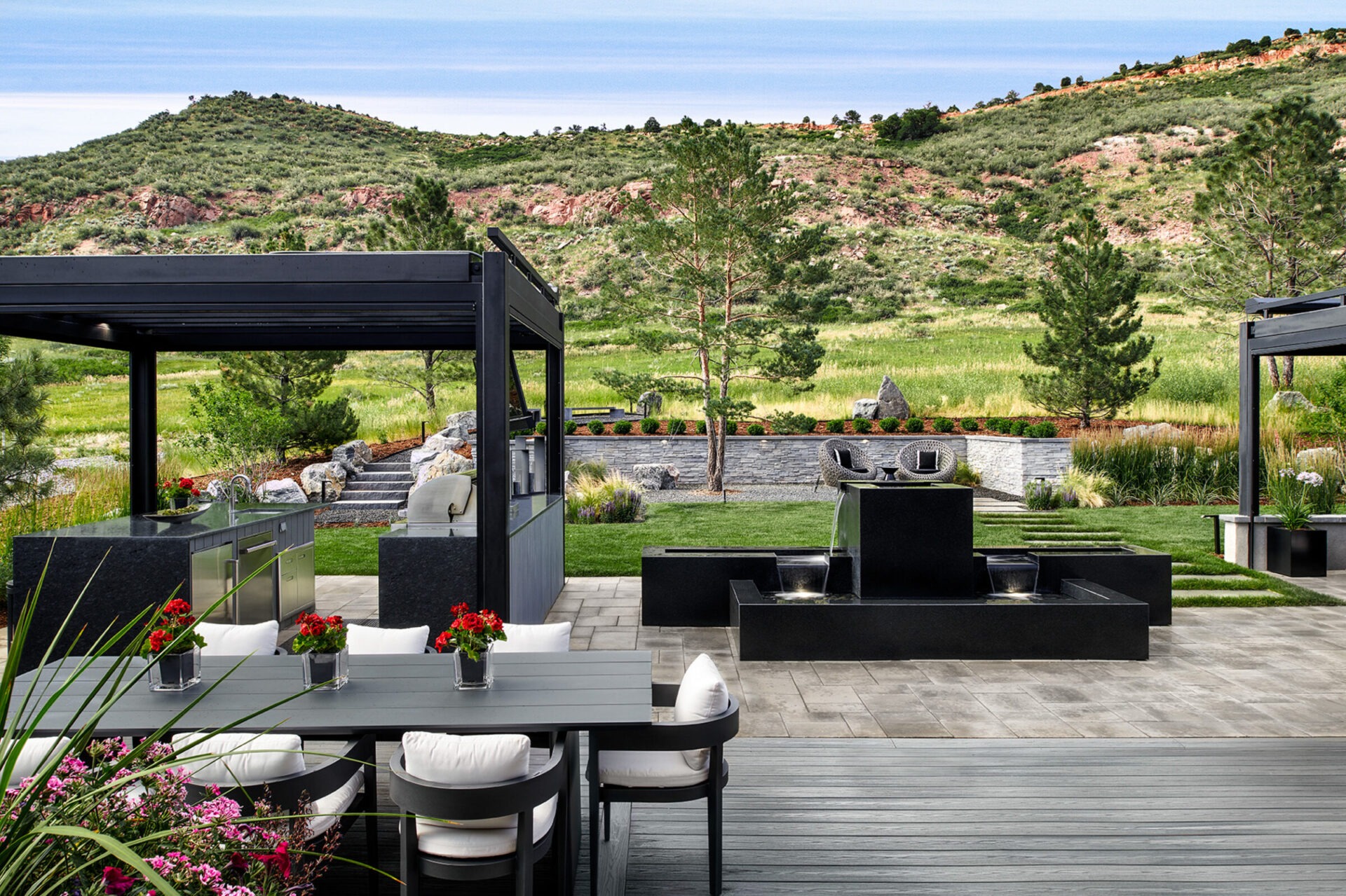 Modern outdoor kitchen with seating, a fountain, and vibrant flowers. Scenic hills and greenery provide a natural backdrop, creating a serene atmosphere.