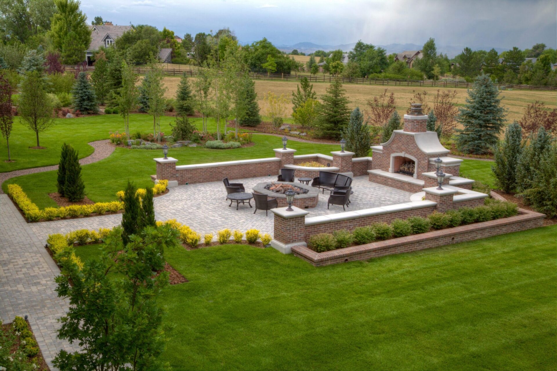 Spacious landscaped garden with paved patio, outdoor fireplace, and seating area, surrounded by lush greenery and fields. Houses visible in the background.