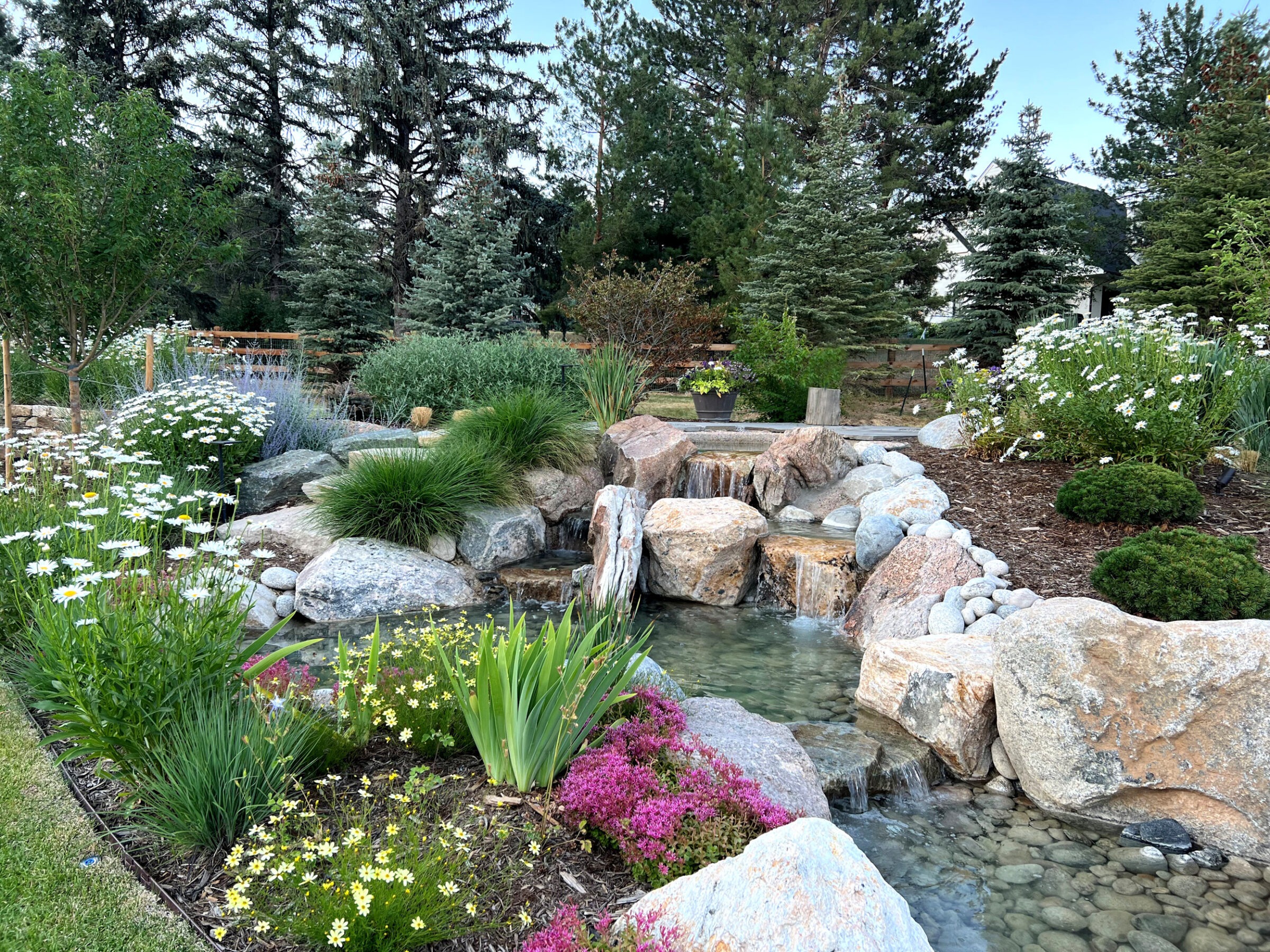 A lush garden featuring a small waterfall with vibrant flowers, lush greenery, and tall trees in the peaceful background.