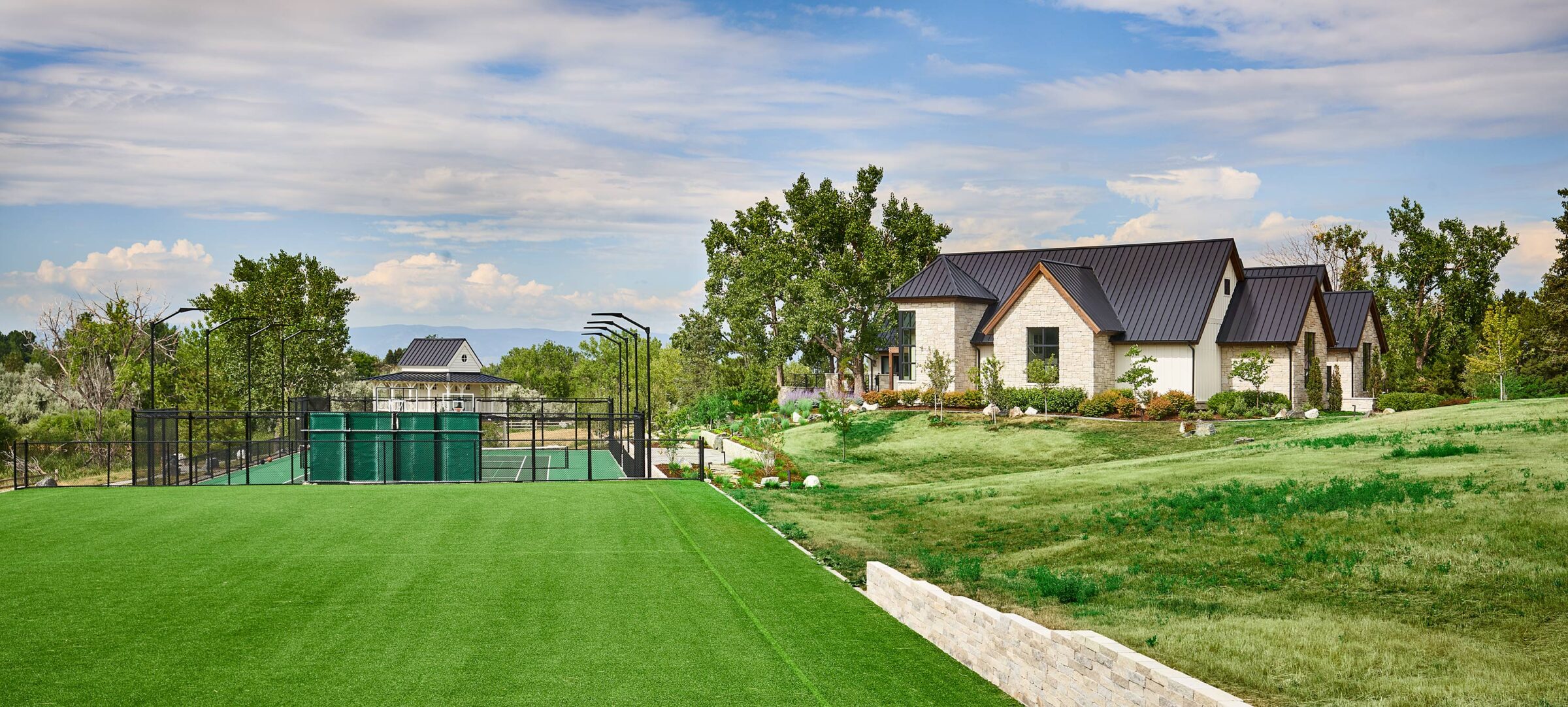 A modern house with dark roof, lush green lawn, and a fenced tennis court surrounded by trees under a partly cloudy sky.