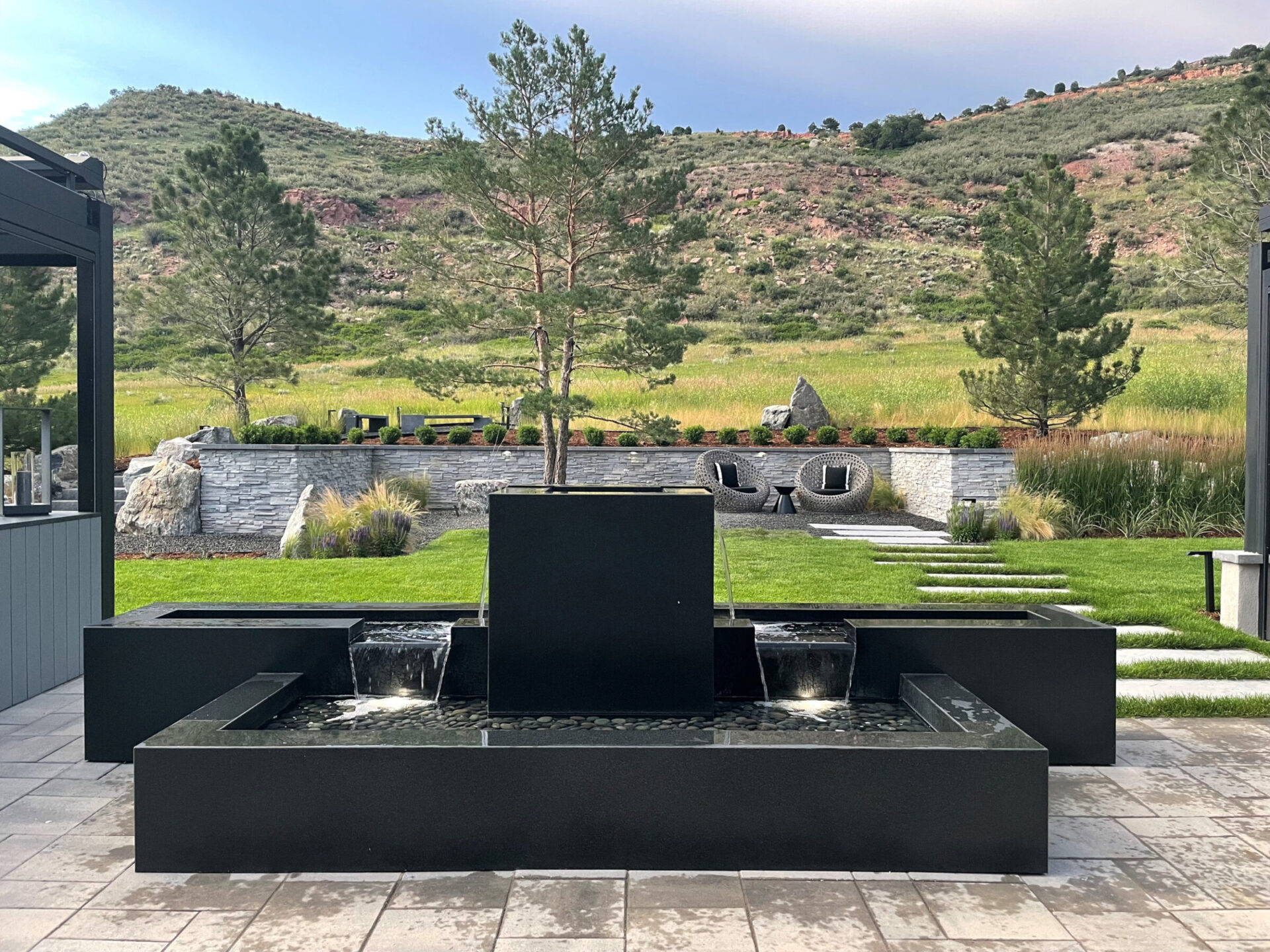 Modern patio with a sleek black fountain, lush green lawn, stepping stones, and scenic hillside view, surrounded by trees and contemporary furniture.