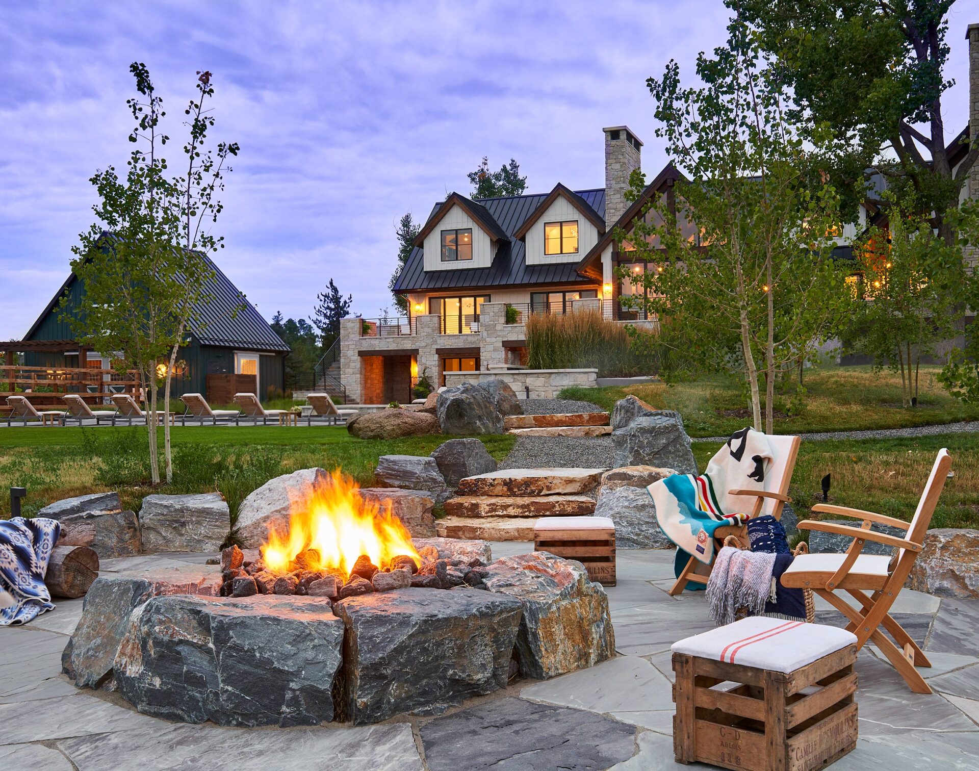 Cozy outdoor fire pit with chairs and blankets on stone patio, surrounded by trees. A warm, inviting house is visible in the background.