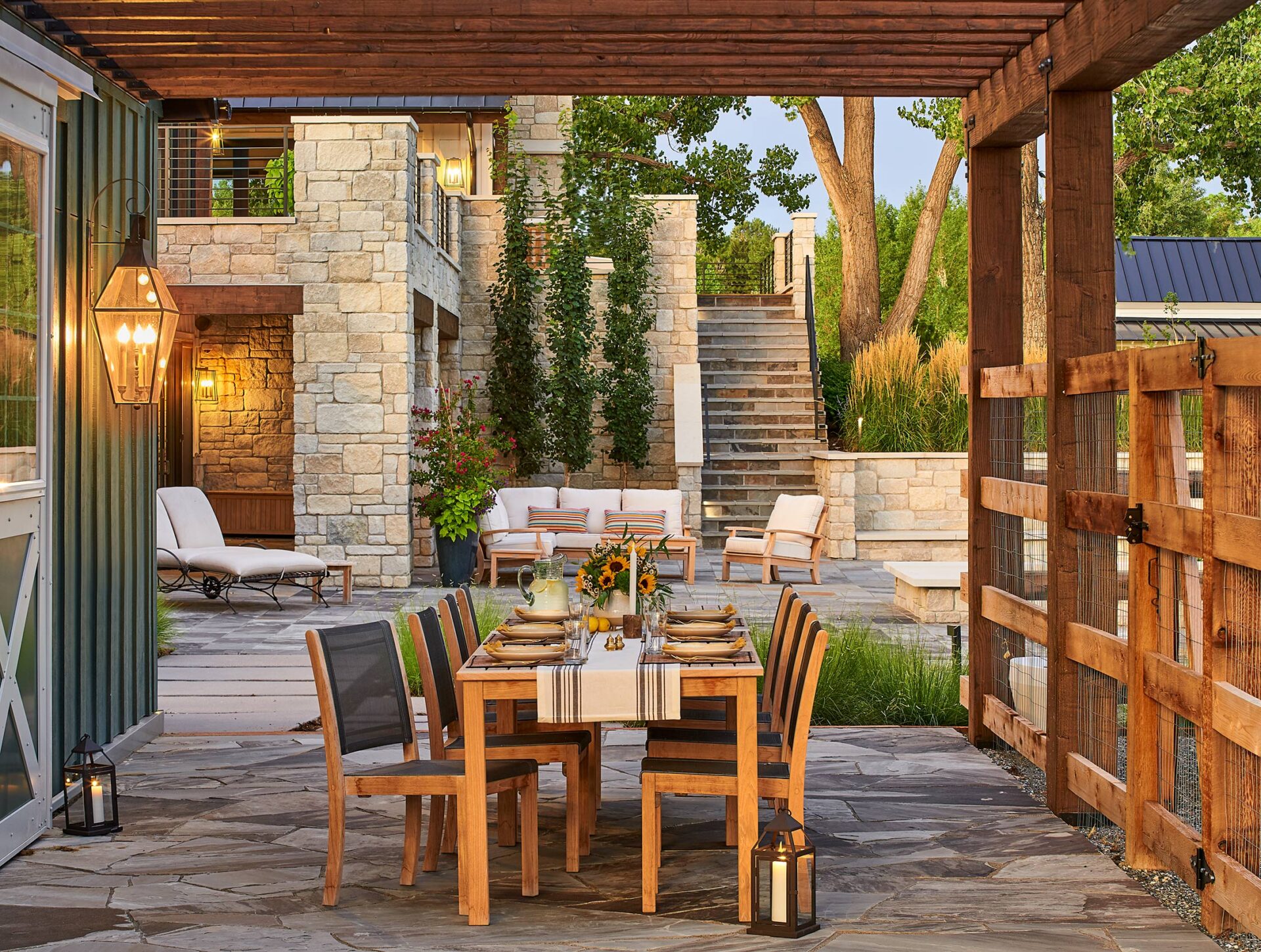 Outdoor patio featuring wooden dining table, chairs set for dinner. Cozy seating area, stone architecture, greenery, soft lighting create a warm, inviting ambiance.