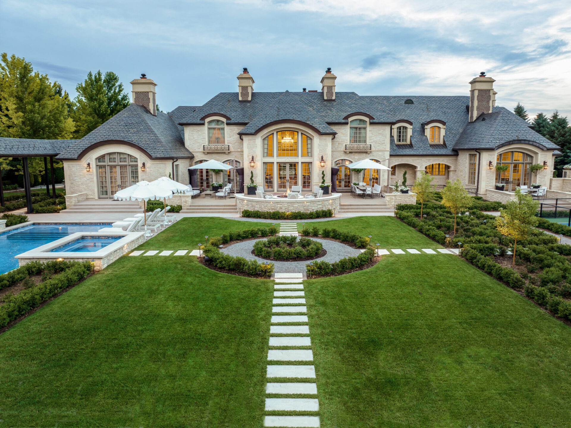 Elegant mansion with large garden, stone pathways, and a pool. Features symmetrical design, multiple chimneys, and outdoor seating under umbrellas. Lush greenery surrounds.