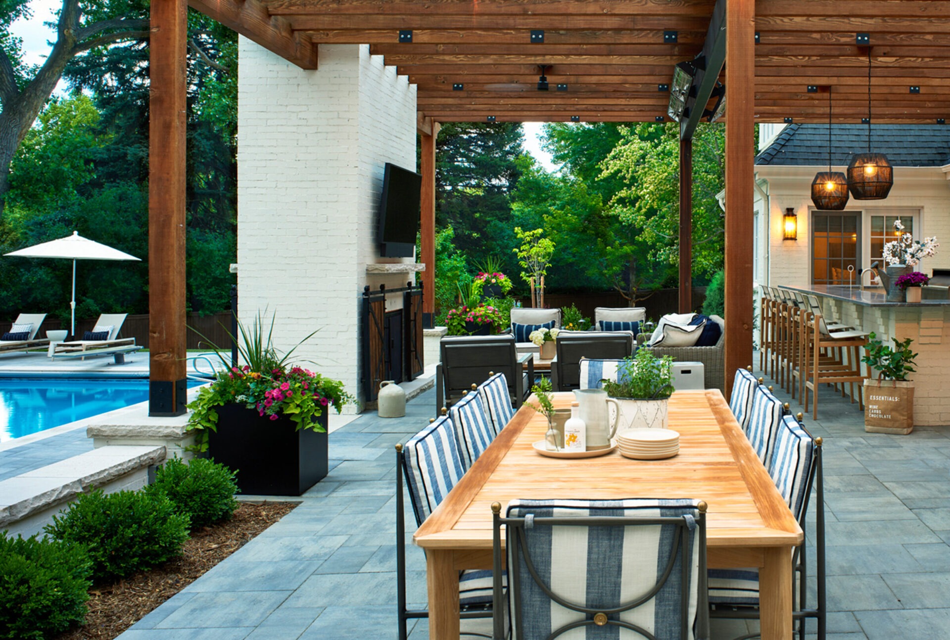 Outdoor patio with wooden pergola, dining table, bar counter, and lounge chairs by a pool. Surrounded by greenery and decorative plants.