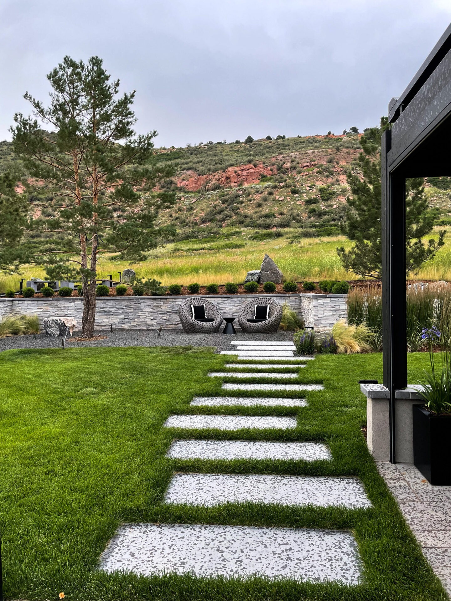 Stepping stone path leads through a landscaped garden with modern seating and scenic hill backdrop. Lush greenery and stone elements create a serene atmosphere.