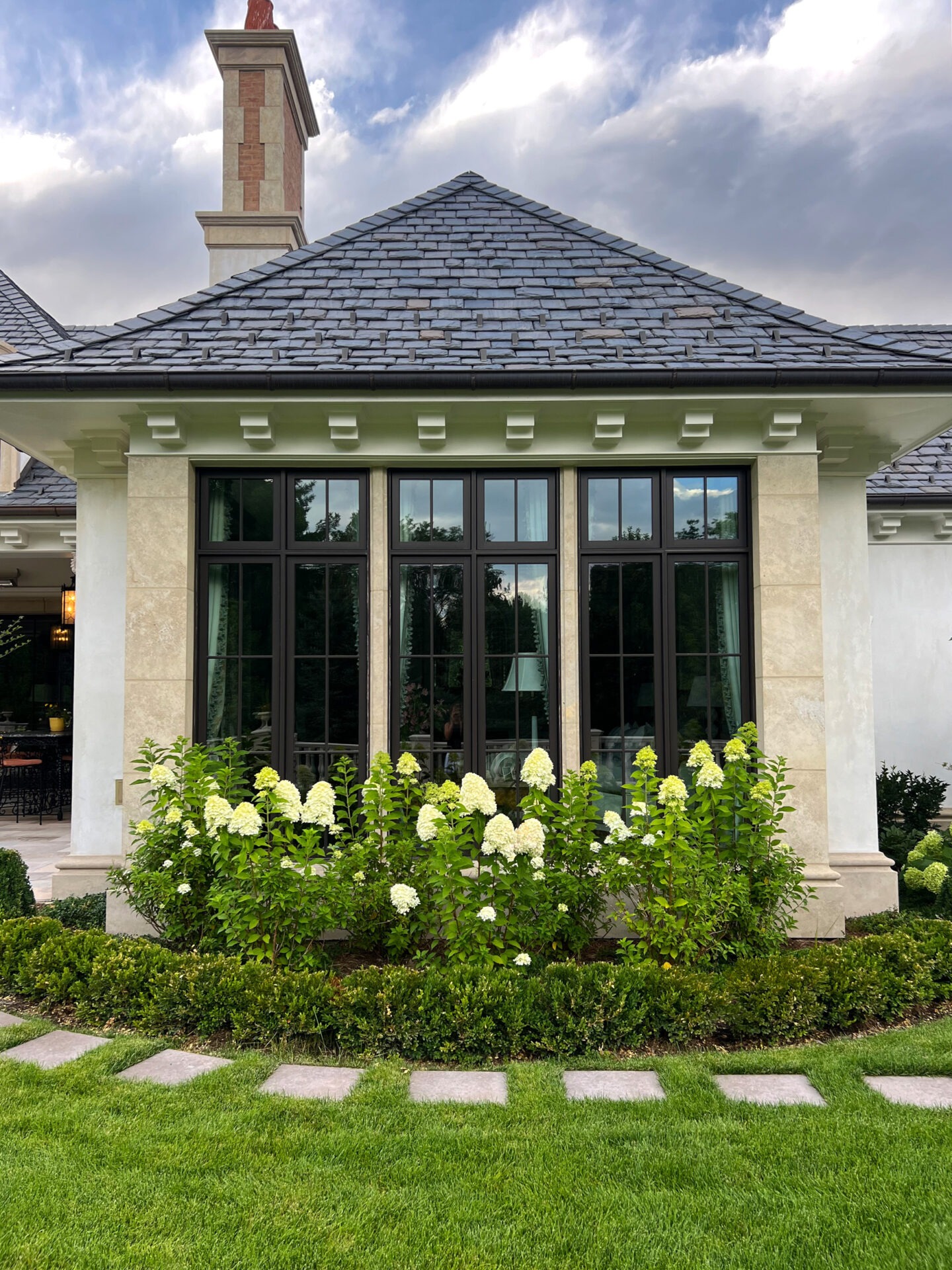 Elegant house exterior with large windows, slate roof, and lush garden featuring hydrangeas. Stone pathway and chimney complement the serene setting.