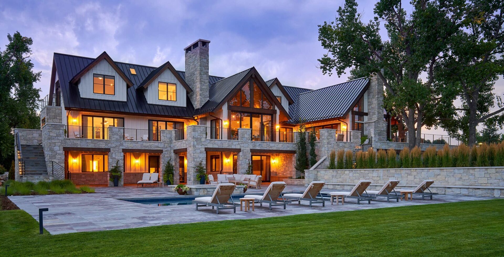 A large, luxurious stone house with multiple gables, lit warmly, surrounded by trees, features a patio and pool with lounge chairs.