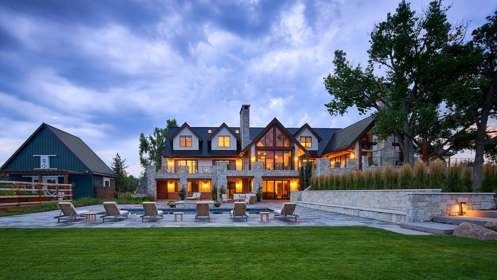 Large, illuminated country house with slate roof, surrounded by trees. Stone terrace features lounge chairs and manicured lawn under a dramatic evening sky.