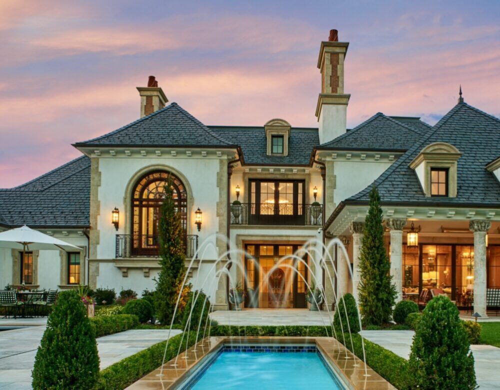 Luxurious mansion with elegant architecture, featuring a fountain, pool, and manicured gardens under a colorful sunset sky. No people present.