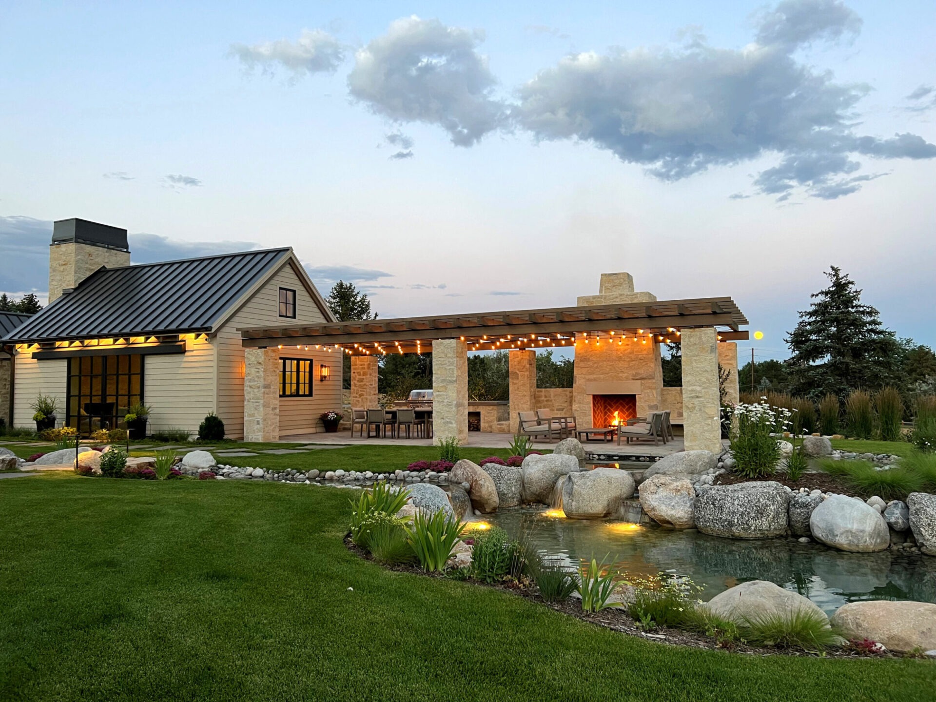 A cozy backyard with a lit pergola, modern house, pond, and landscaped garden under a twilight sky with scattered clouds.