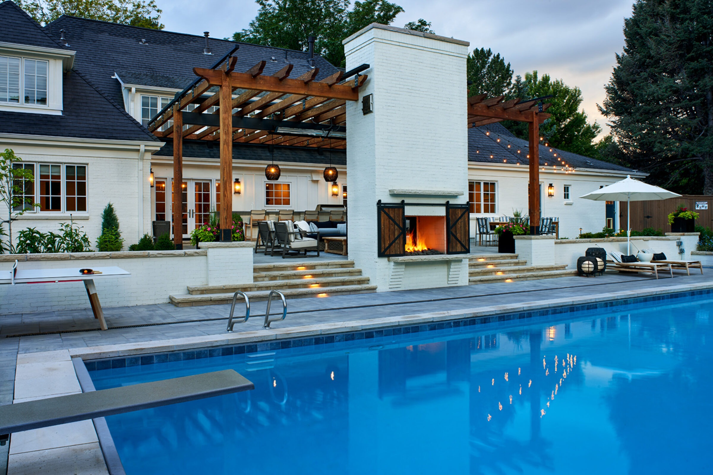 A luxurious backyard features a modern patio with a fireplace, string lights, seating area, ping pong table, and a swimming pool.