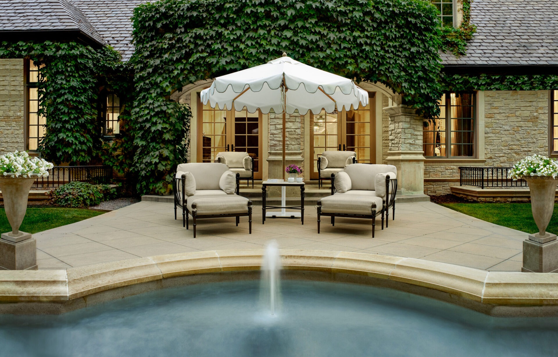 Elegant patio with white umbrella, beige chairs, and stone facade. Ivy climbs walls around glass doors. Circular fountain in foreground.