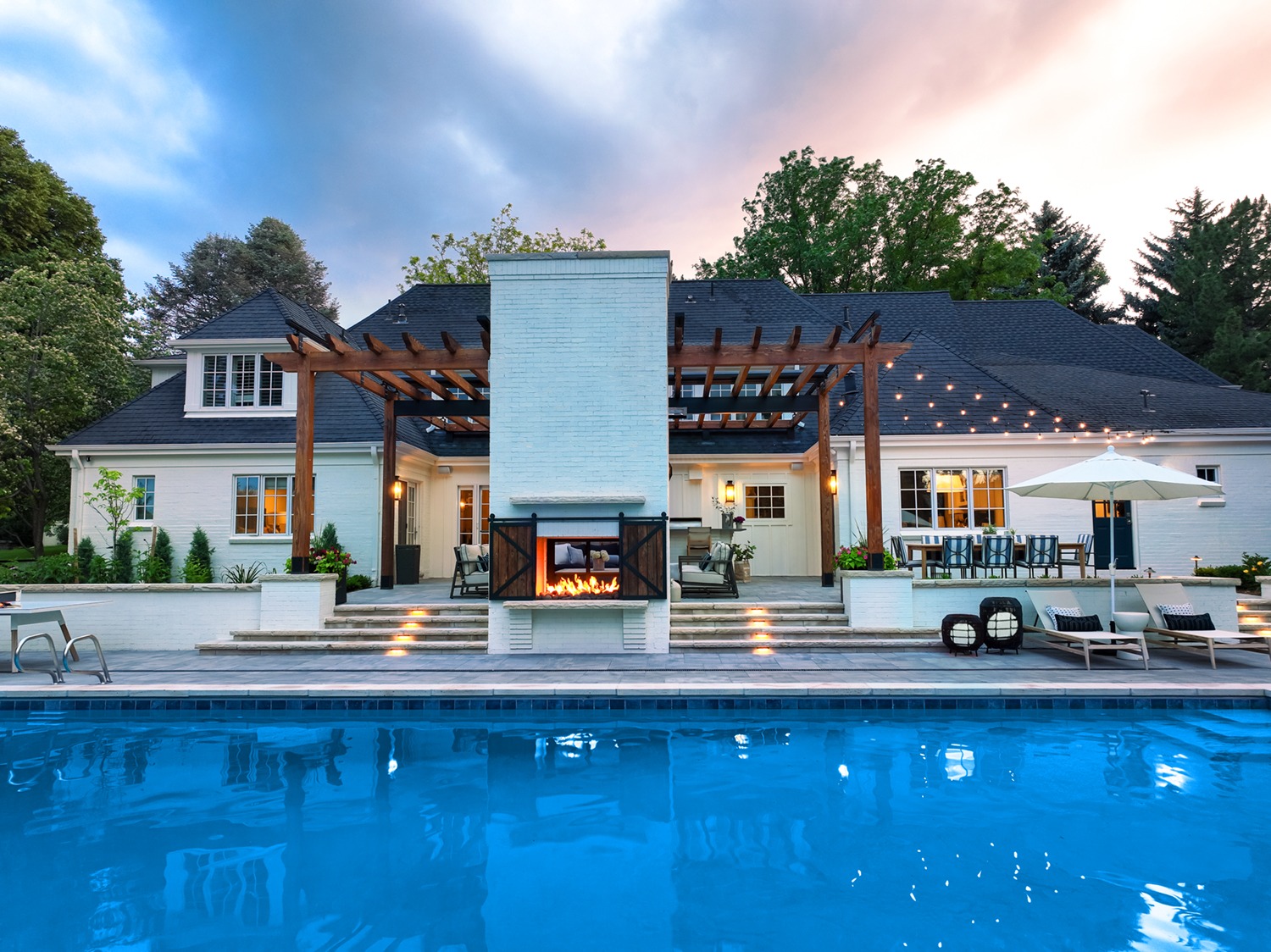 A modern house with glowing outdoor fireplace, pergola, and poolside lounge area, surrounded by lush trees and serene sky at dusk.