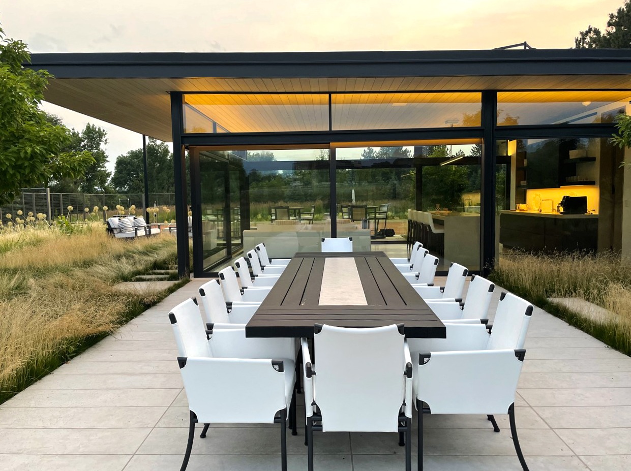 Modern glass house with outdoor dining table, surrounded by grass and trees. Warm interior lighting creates a cozy, inviting atmosphere.