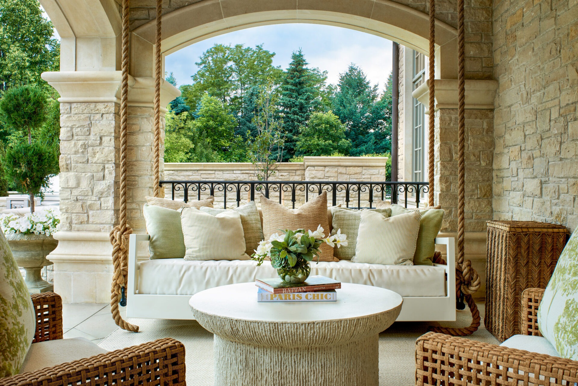 Chic outdoor patio with a swinging couch, cozy cushions, and a round table. Surrounded by greenery and a stone-finish exterior.