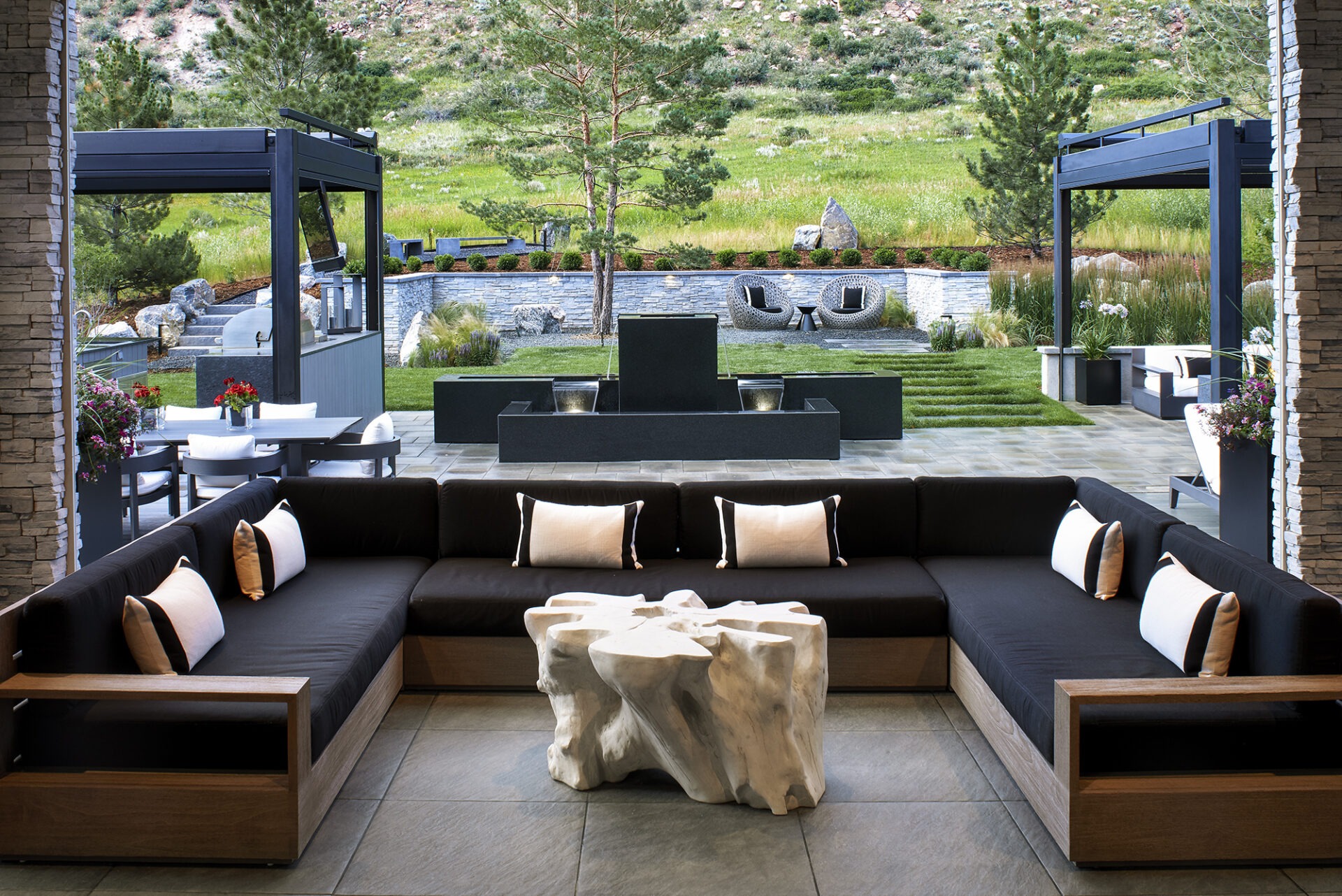 Modern outdoor seating with black cushions, unique table, manicured lawn, and decorative fountains, surrounded by greenery and stone accents under clear skies.