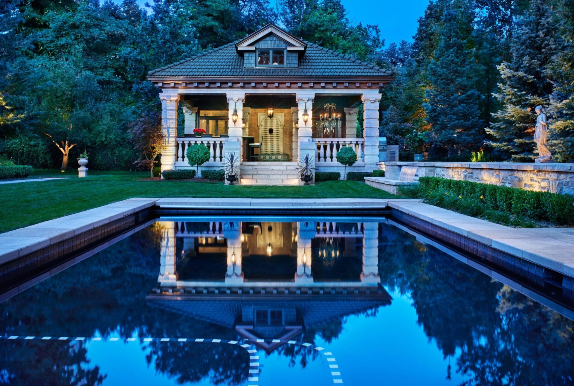 A charming small house with columns, surrounded by greenery, overlooks a reflective swimming pool at twilight, creating an inviting, serene atmosphere.