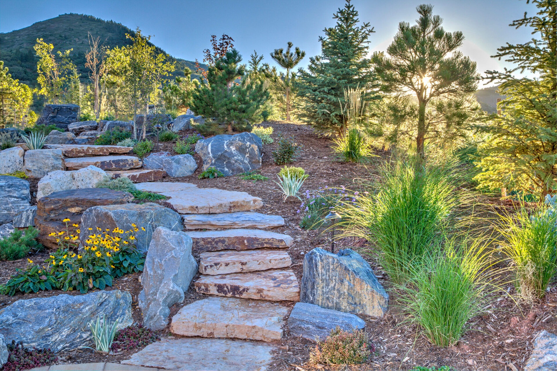 A serene garden landscape features stone steps, lush greenery, and vibrant flowers against a backdrop of a sunlit mountain and clear sky.