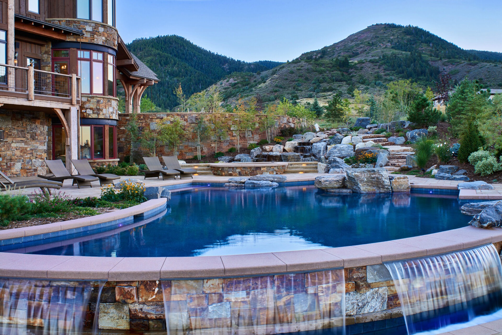 A luxurious mountain retreat features an infinity pool with cascading waterfall, stone facade, and lush landscaping against serene hillside backdrop.