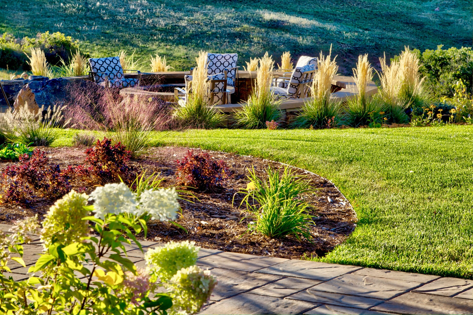 A lush garden with blooming flowers, patio seating, and ornamental grasses, set against a backdrop of green hills in sunlight.