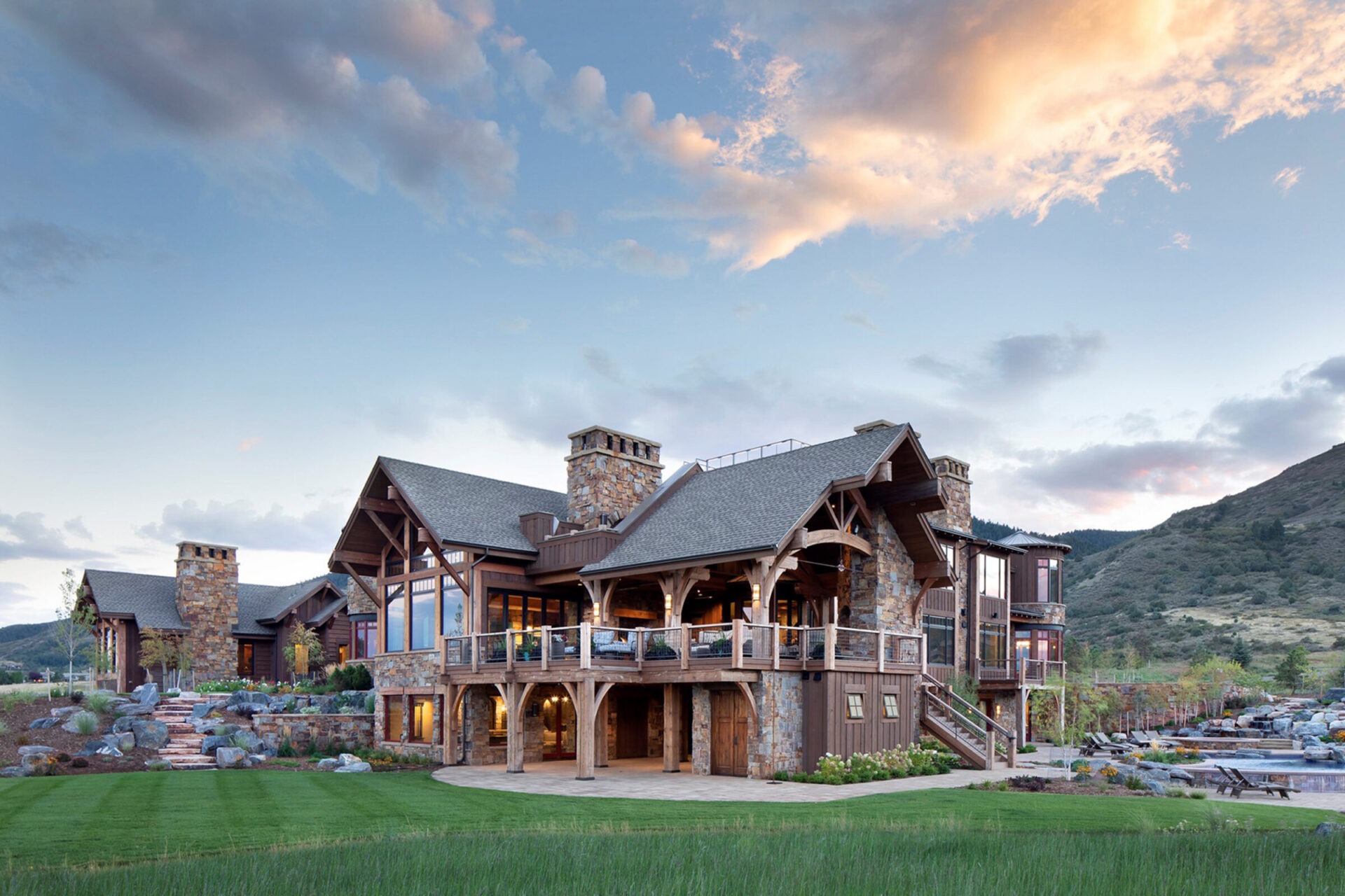 Large rustic house with wooden beams, surrounded by lush greenery and mountains under a partly cloudy sky. Features stone chimneys and landscaped garden.