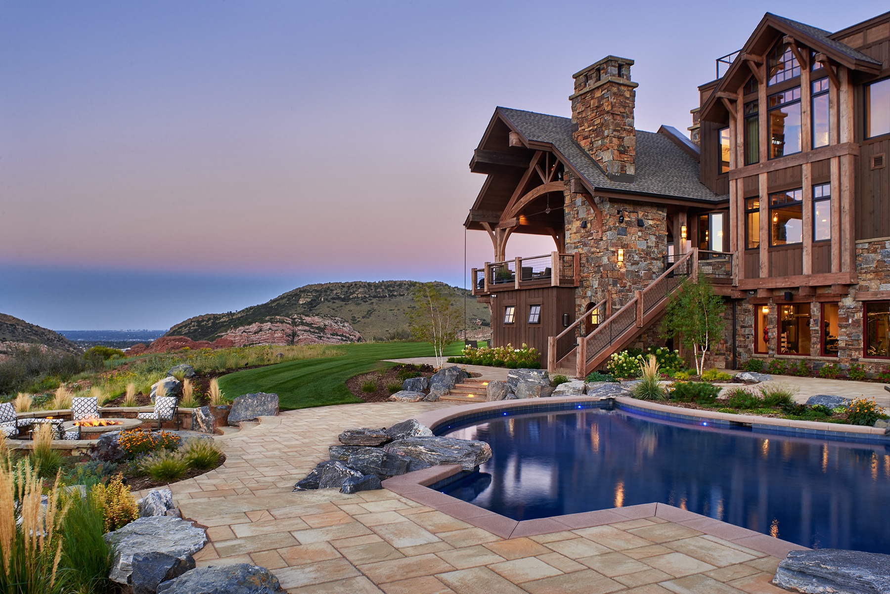 A rustic stone house features a pool, fire pit, and scenic mountain view during twilight, surrounded by landscaped gardens and paved pathways.