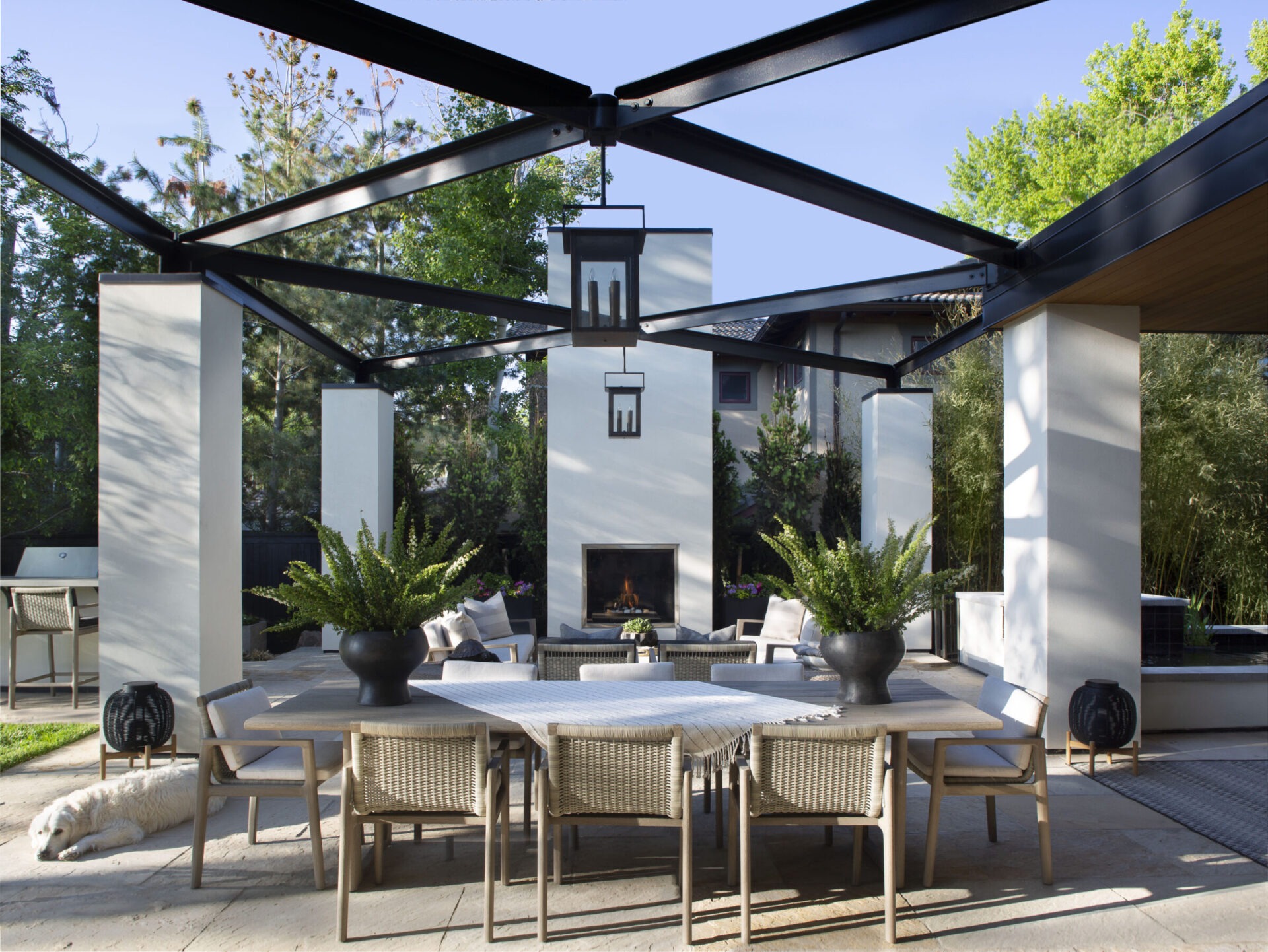 Modern outdoor patio with a dining table, chairs, fireplace, and plants. A dog rests in the shade. Surrounded by greenery and trees.