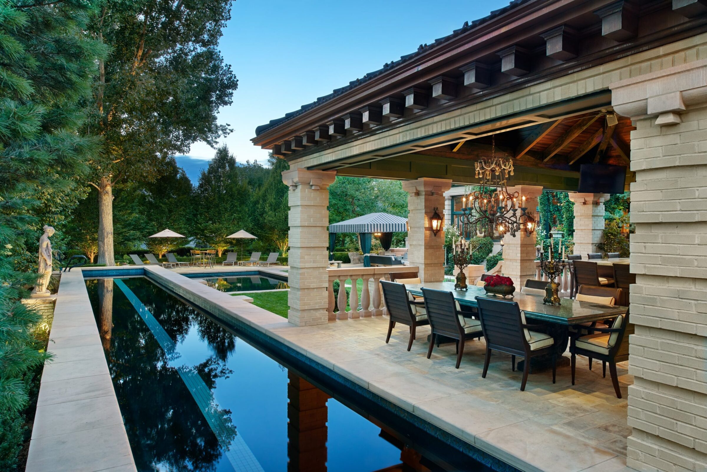 Elegant outdoor patio with pool, dining area, chandeliers, and garden. Columned structure, surrounded by lush greenery and a peaceful atmosphere.