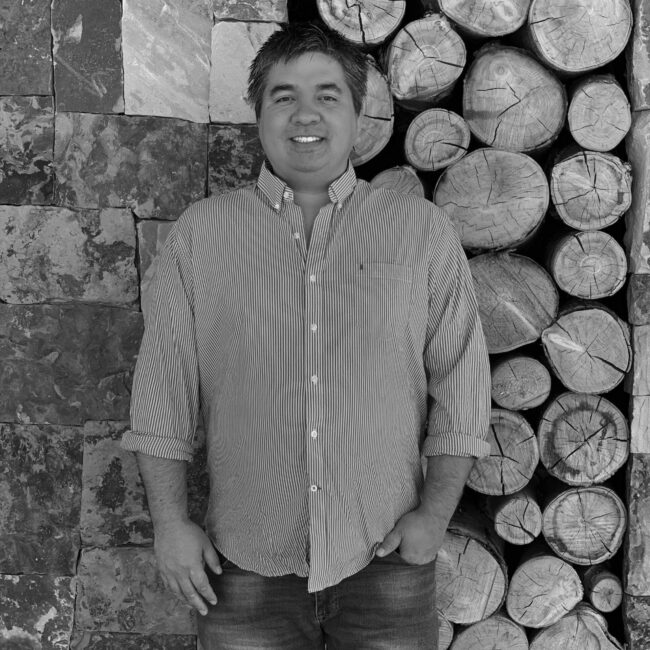 A person stands smiling, wearing a striped shirt beside stacked logs and a textured stone wall in a serene environment.