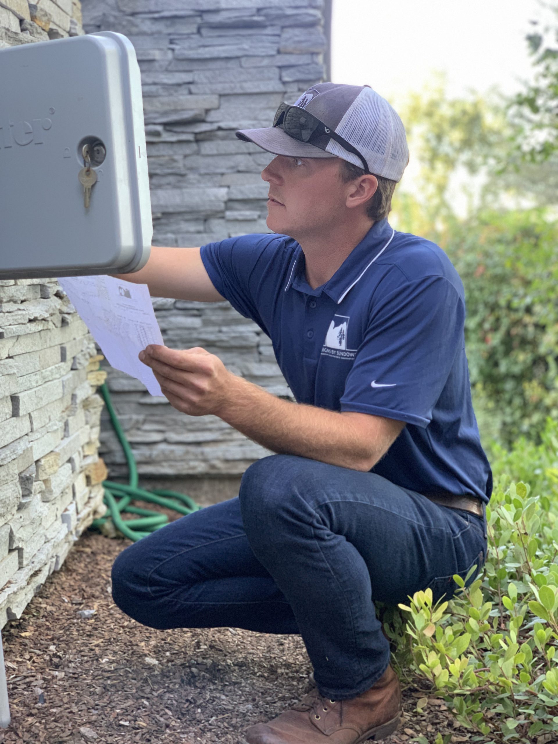 A person in casual attire inspects an outdoor electrical box, holding papers, surrounded by greenery and stacked stone wall.