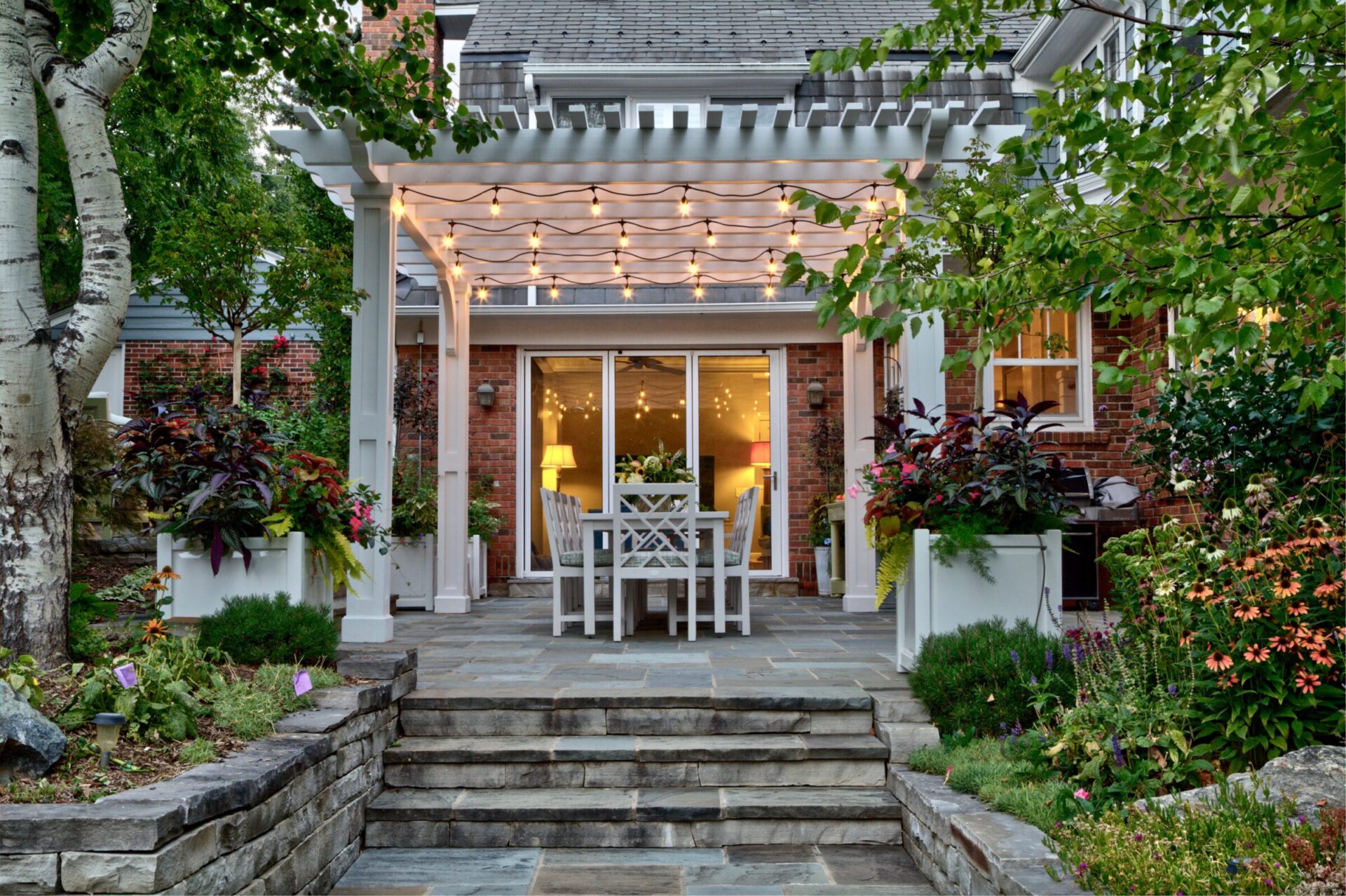 Cozy backyard with a white pergola and string lights, surrounded by lush plants and brick walls. Inviting outdoor dining area visible.