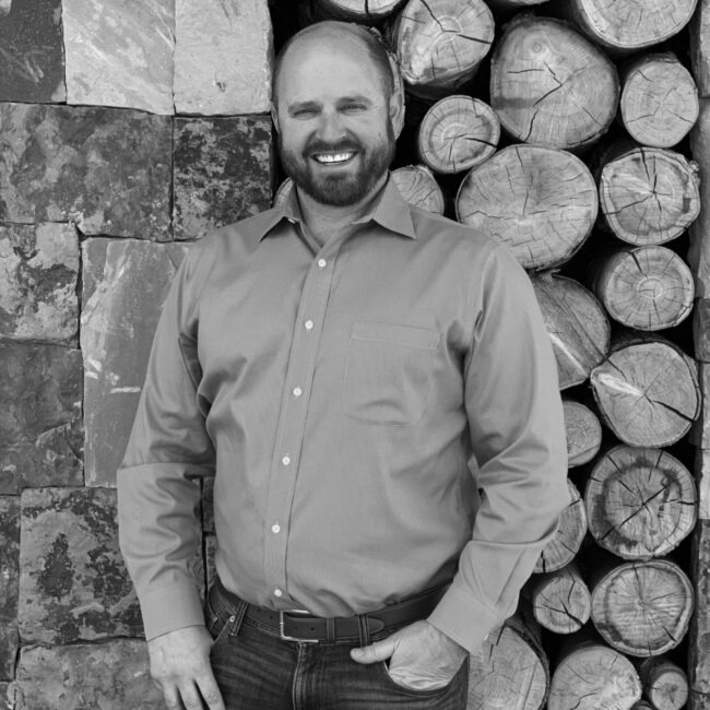 A person in a button-up shirt stands smiling beside a stack of firewood and a stone wall, in a black and white photograph.