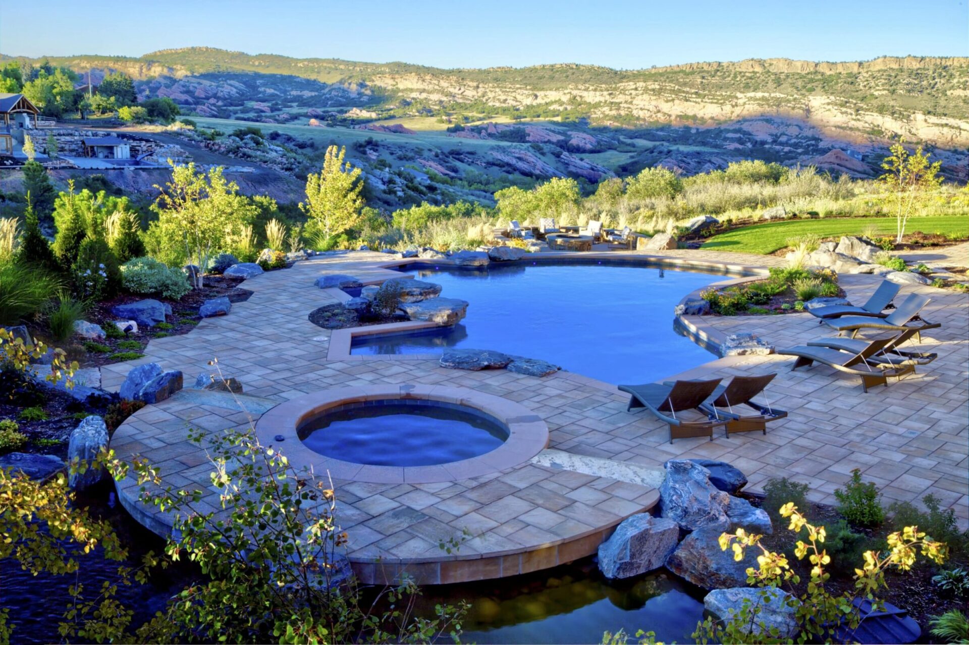 Luxurious pool and hot tub set against a scenic backdrop of rolling hills, surrounded by lush greenery and several lounge chairs.