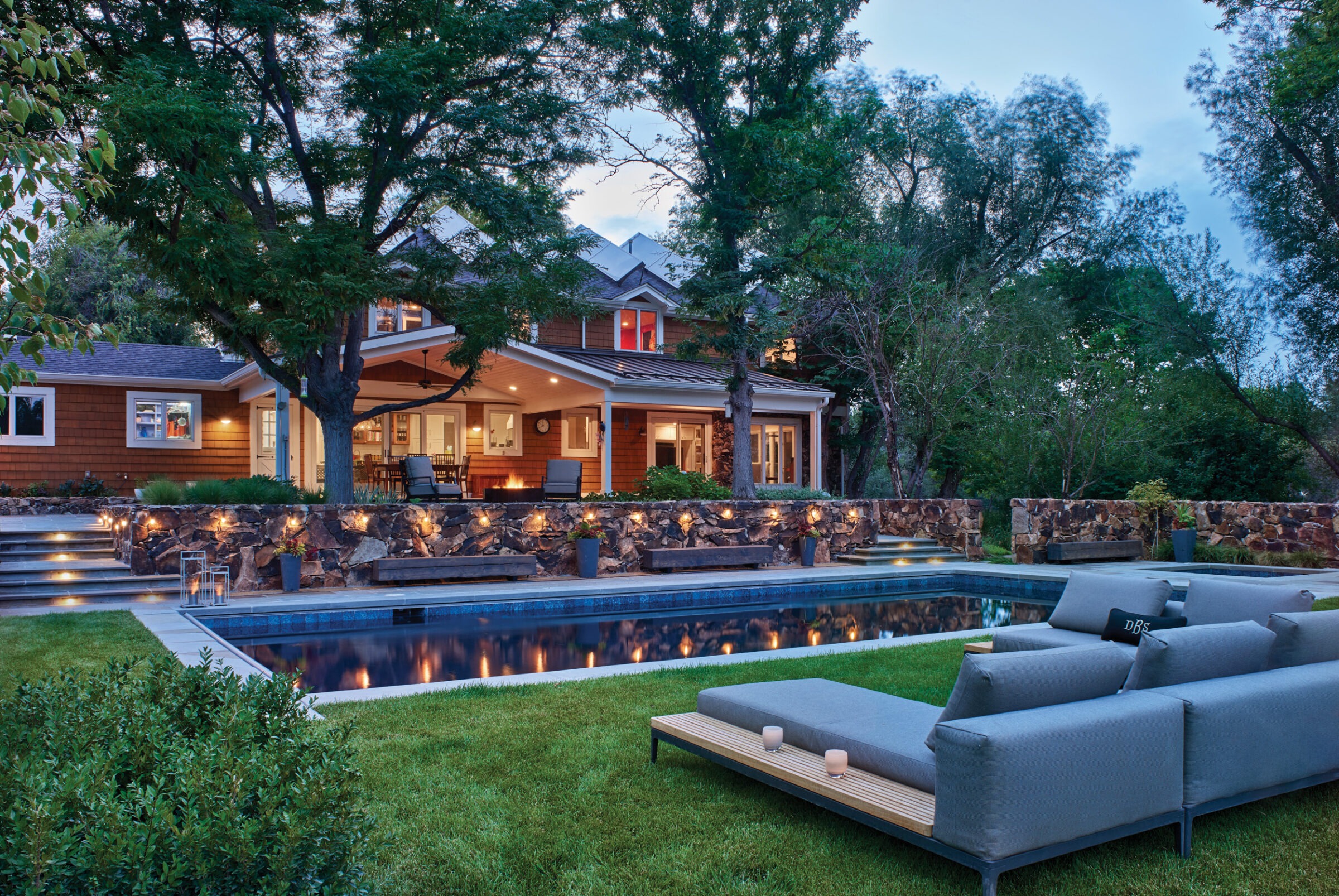 A modern house with open patio, surrounded by trees. In the foreground, a pool and outdoor seating on a grassy area.