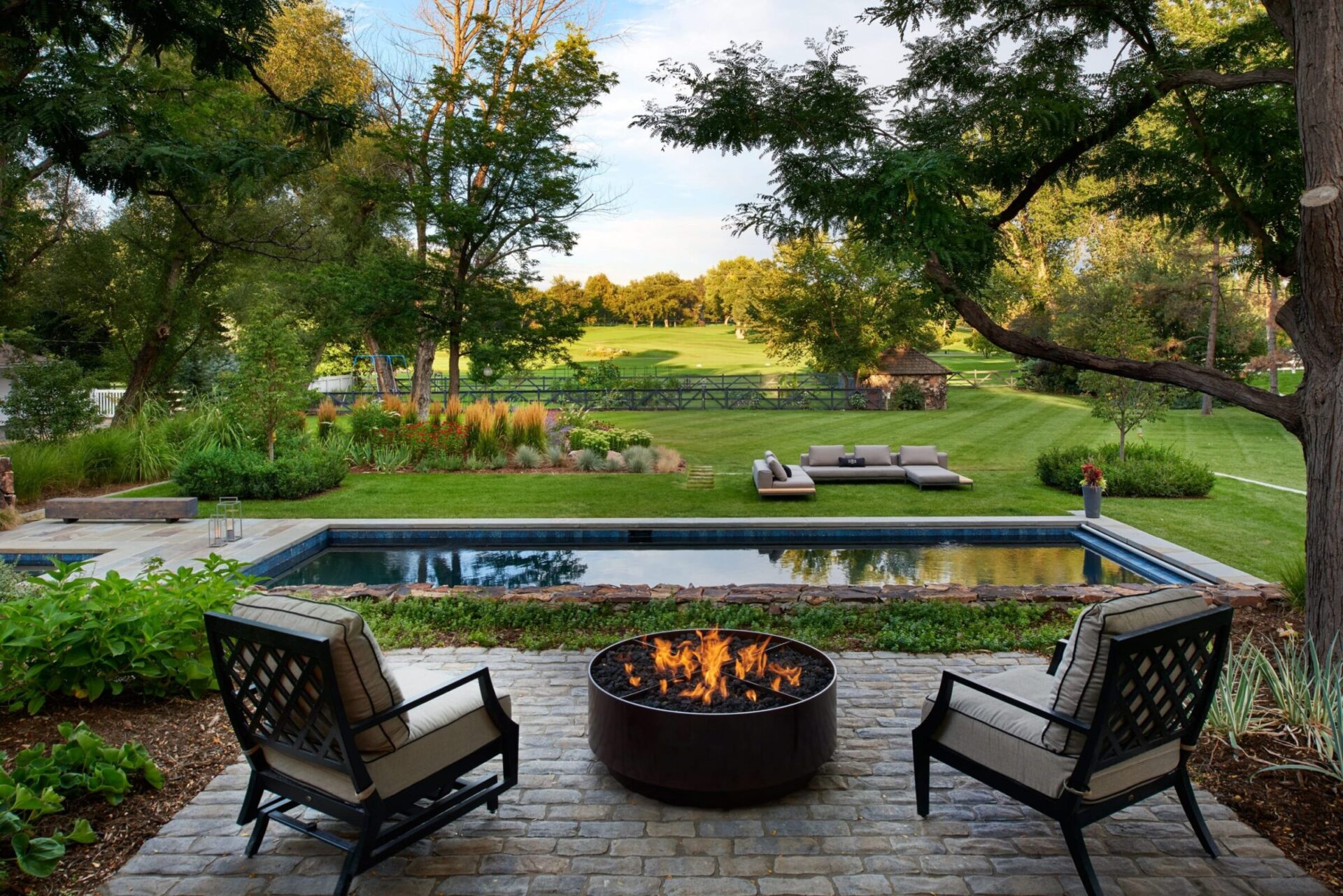 A tranquil backyard scene with a fire pit, pool, and lush garden, featuring outdoor seating overlooking a grassy area and trees.