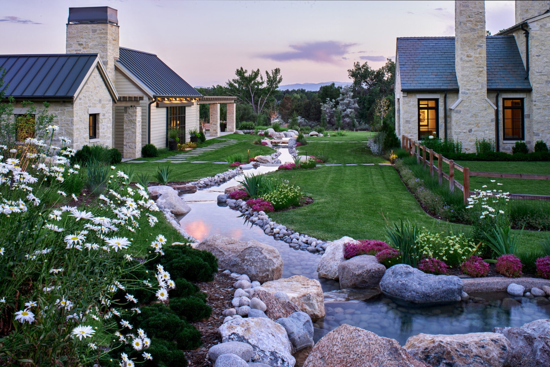 Stone houses with pitched roofs by a landscaped garden, featuring a stream, boulders, flowers, and trees under a twilight sky.