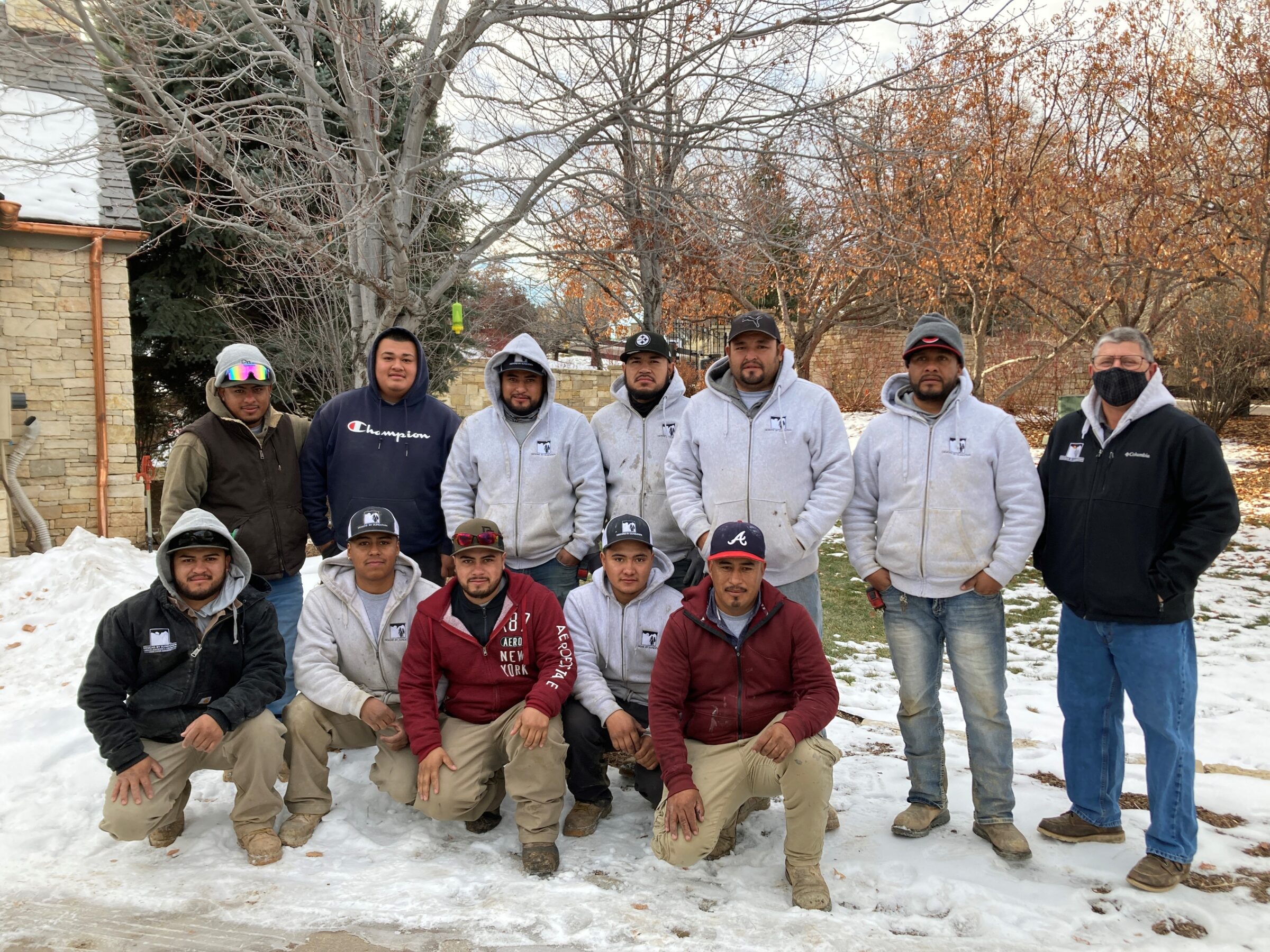 Twelve people in winter clothing pose outdoors on a snowy day. They are standing and kneeling in front of a tree and brick building.