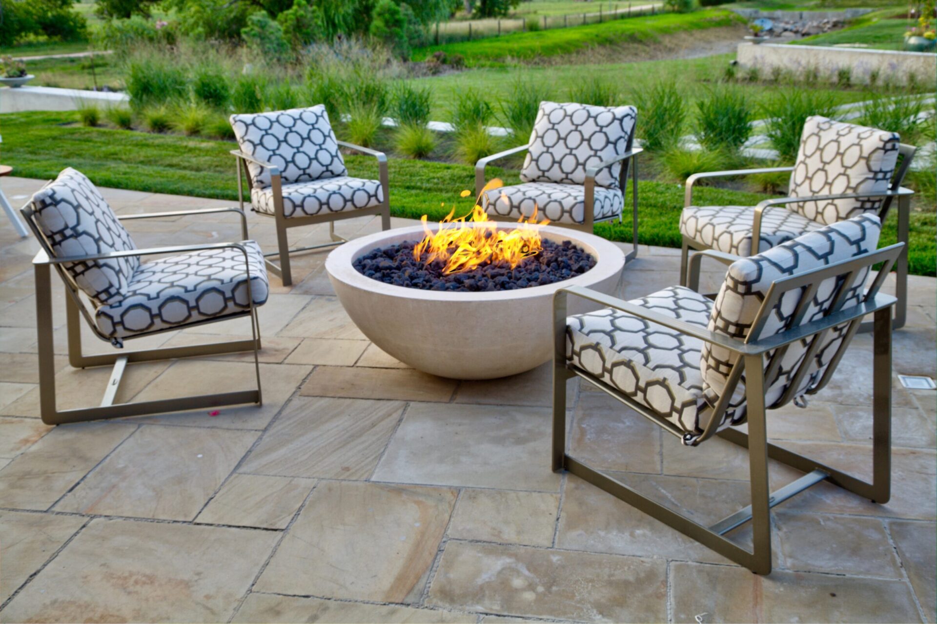 Five patterned chairs surround a lit fire pit on a stone patio, with green grass and trees visible in the background.