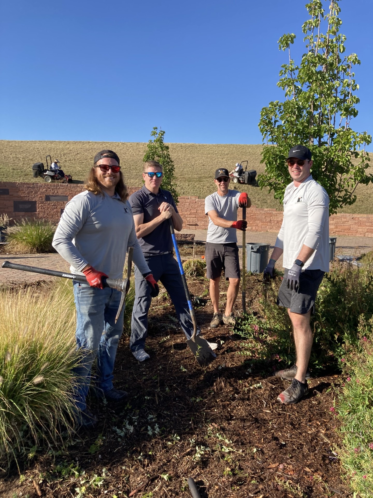 Four people with gardening tools working in a garden under clear sky, with a mowed lawn in the background.