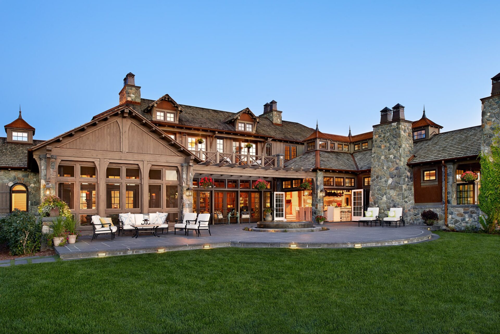 Large rustic house with stone chimneys, wooden exterior, and outdoor seating on a patio. Well-maintained lawn in the foreground, clear sky above.