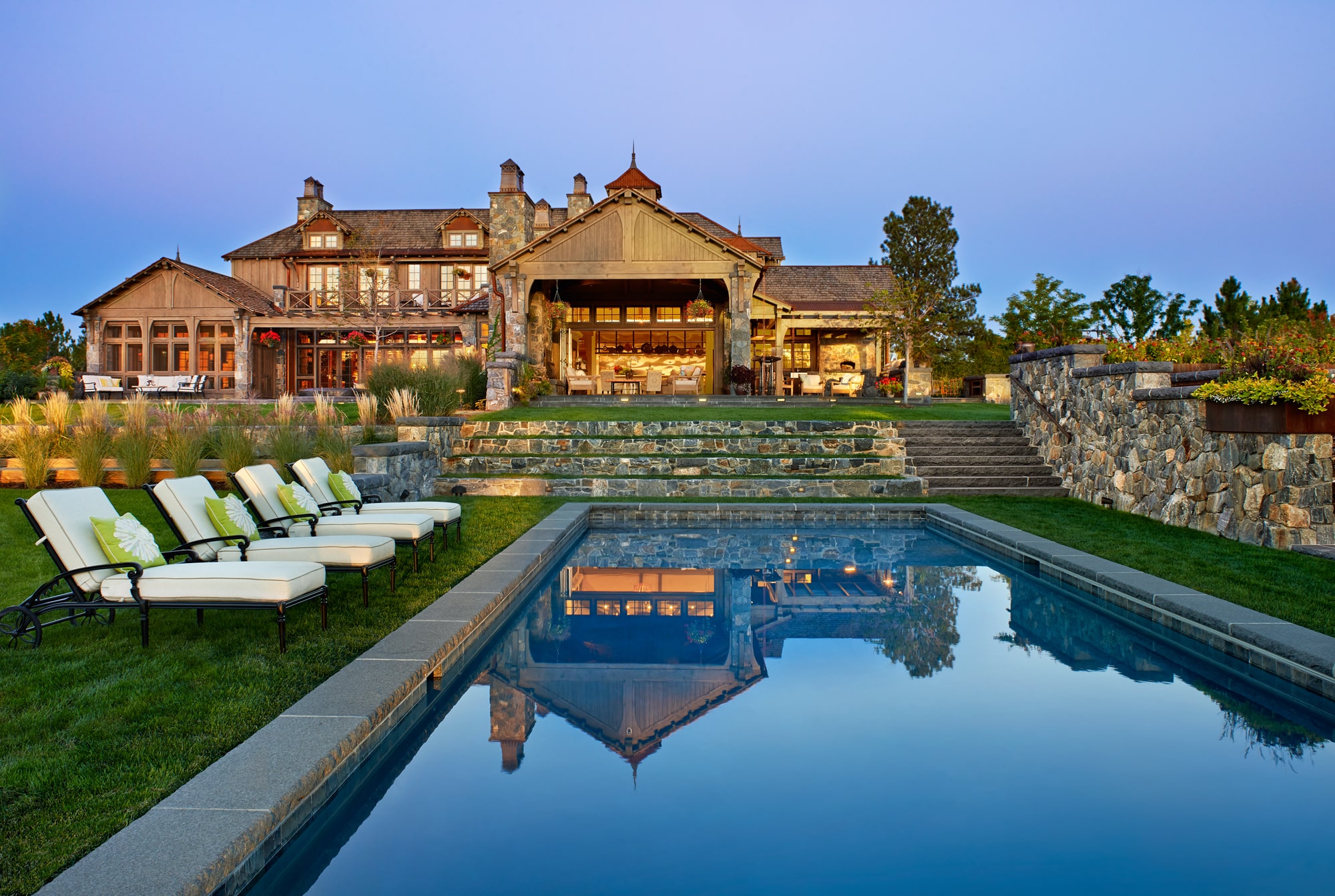 Rustic mansion with stone facade reflected in a serene pool, surrounded by lush landscaping and lined with cushioned lounge chairs.