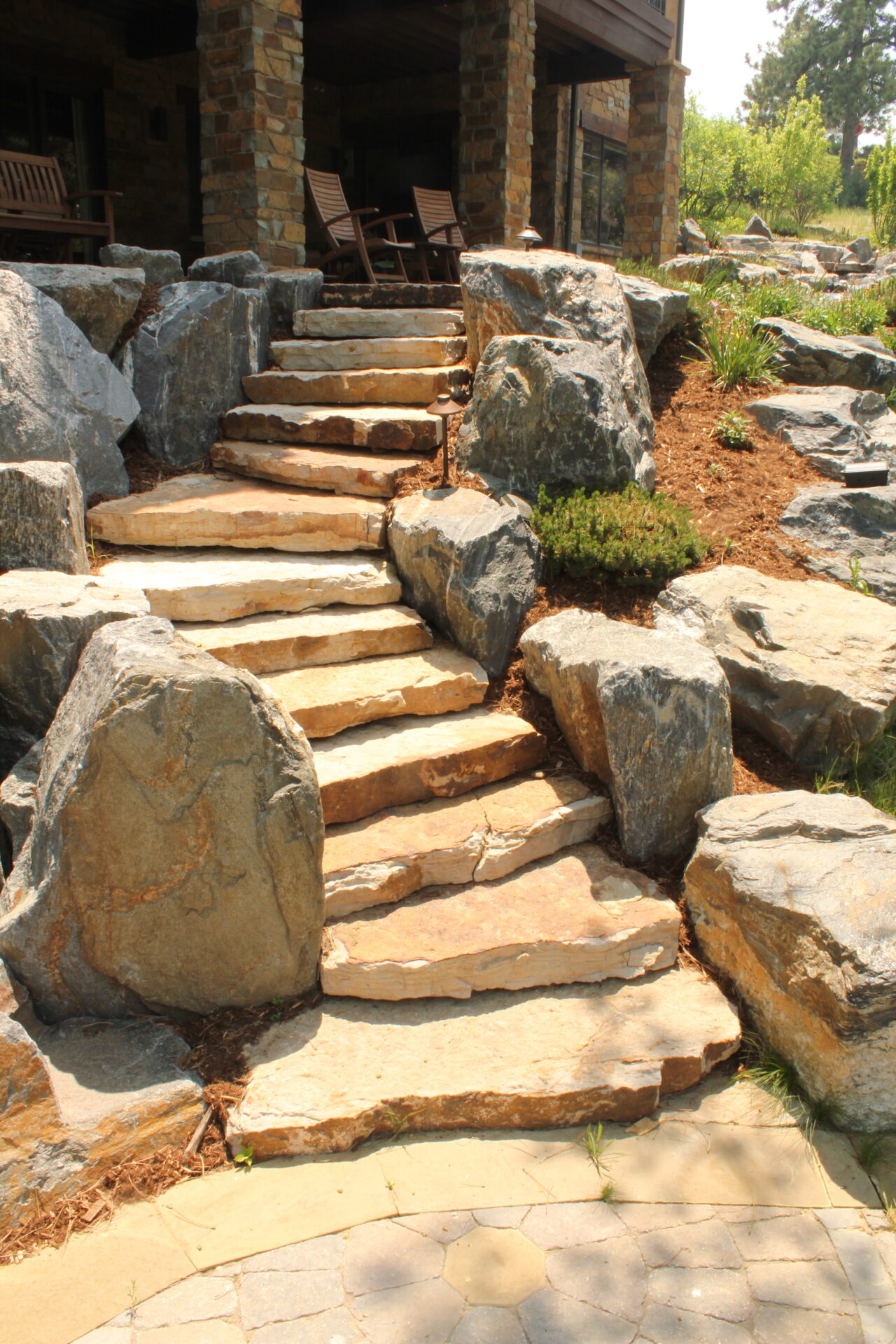 Stone steps lead to a rustic patio with rockers, surrounded by large boulders and greenery, creating a natural, serene atmosphere.