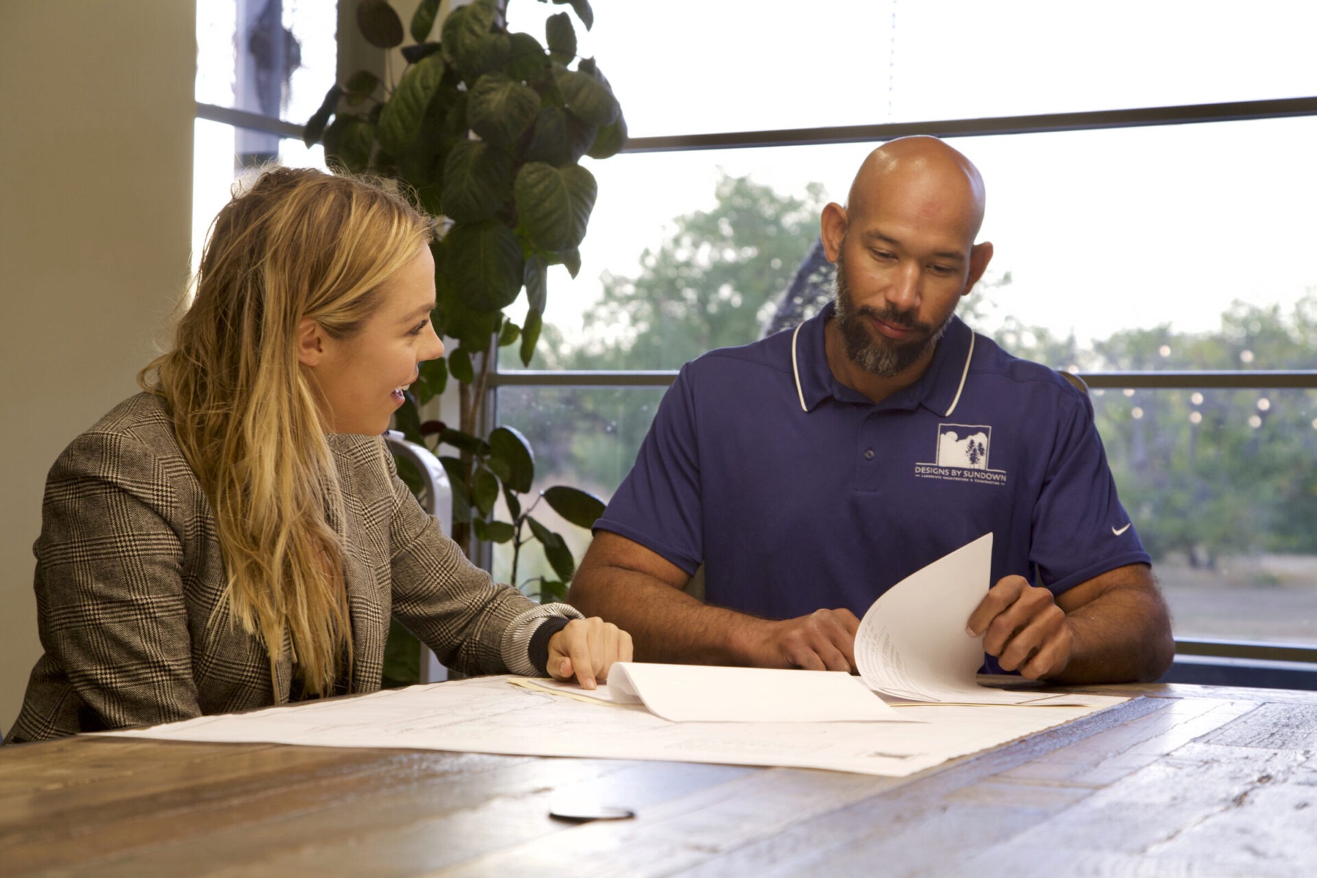 Two people discussing documents at a table indoors; a plant and a window are visible in the background.