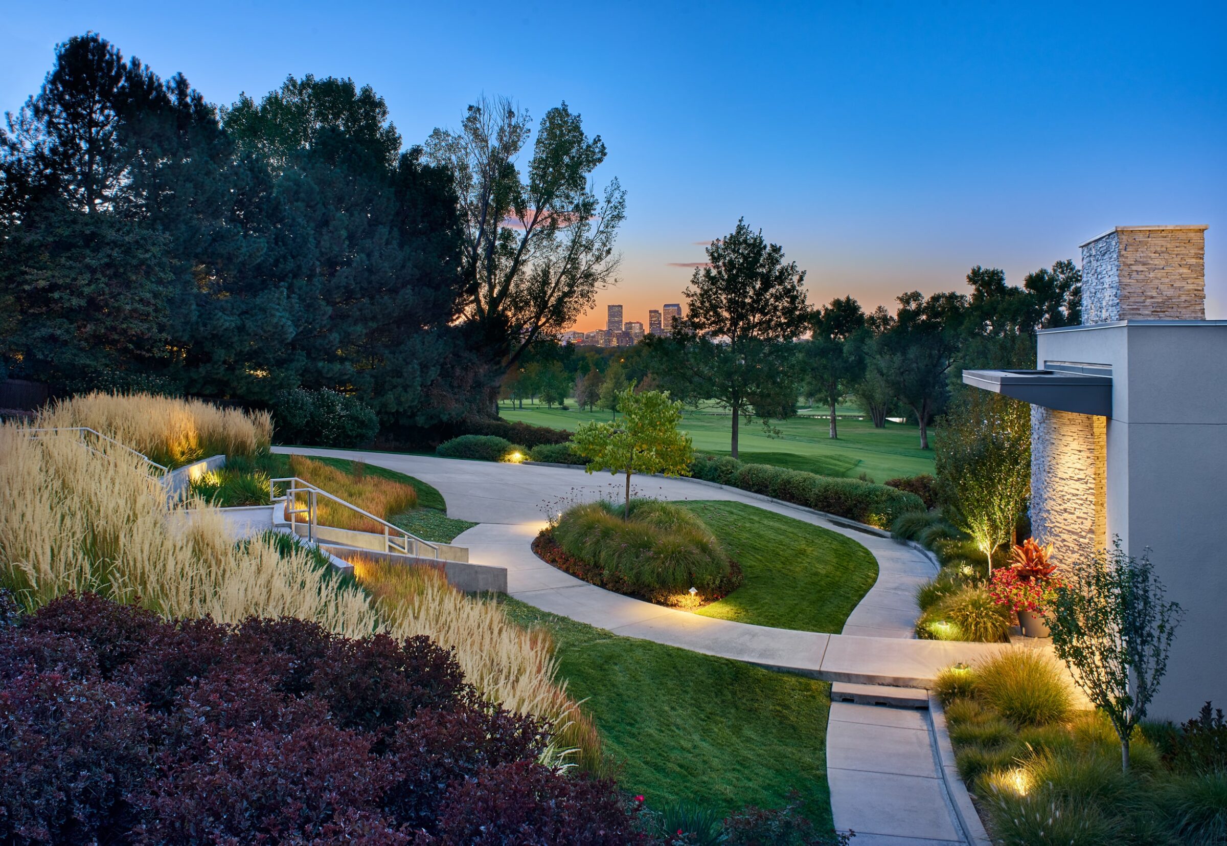 Modern building with garden path, illuminated at dusk. Downtown buildings visible in background, surrounded by trees and greenery. Peaceful, scenic atmosphere.