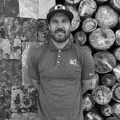 A person wearing a cap stands against a wall of stacked logs and stone blocks, wearing a polo shirt. Black and white image.