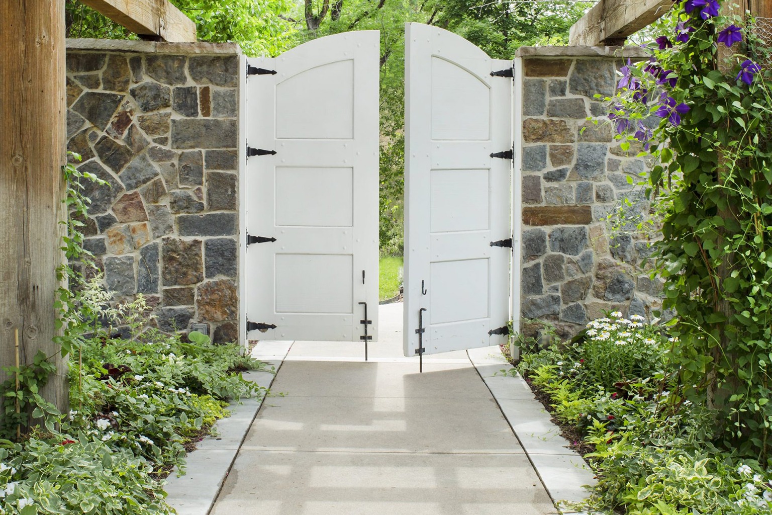 White wooden gate open between stone walls, surrounded by lush greenery and flowering plants, leading to a bright, sunny path ahead.